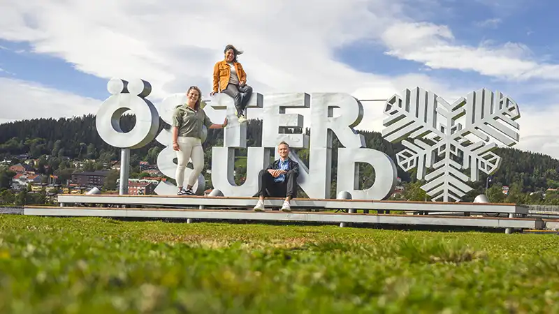 Tre unga människor sitter på den stora Östersundsskylten med Frösön i bakgrunden