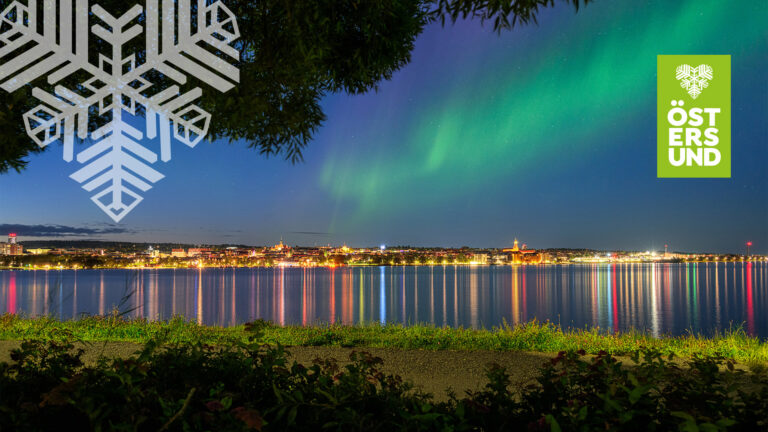 Vy mot Östersunds upplysta stadskärna med ett norrsken i himlen.