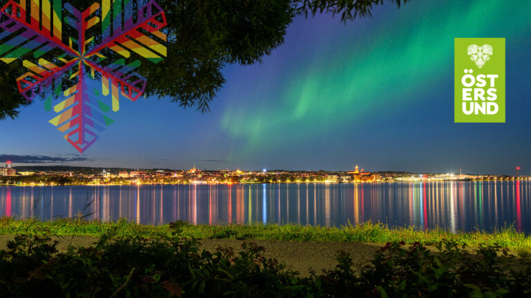Vy mot Östersunds upplysta stadskärna med ett norrsken i himlen.