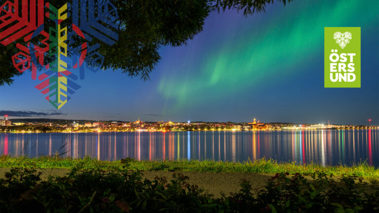 Vy mot Östersunds upplysta stadskärna med ett norrsken i himlen.