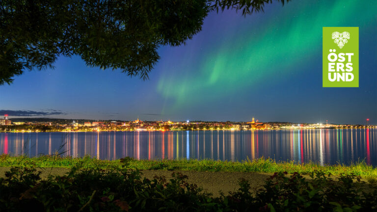 Vy mot Östersunds upplysta stadskärna med ett norrsken i himlen.