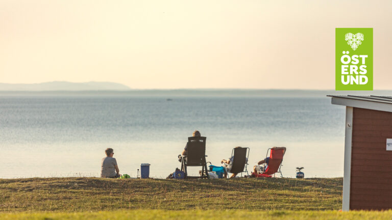 Fyra personer sitter vid strandkanten vid Bynäset och tittar ut mot Storsjöflaket.